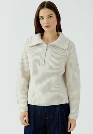 Vrouw met stijlbruin haar die een crème geribde gebreide trui met een grote kraag en rits draagt, gecombineerd met een donkerblauwe broek.