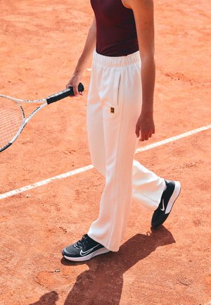 Person in weißen Jogginghosen und schwarzen Nike-Sneakern hält einen Tennisschläger und steht auf einem Sand-Tennisplatz.