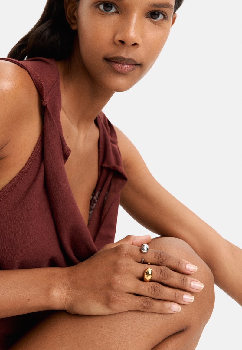 Dwa pierścionki na dłoni: jeden błyszcząca srebrna kula i jeden złoty owal, na tle, które podkreśla teksturę i kolor skóry.