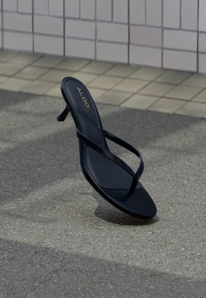Sandale negre cu toc, cu bretele subțiri și branding ALDO, poziționate pe asfalt lângă o pavimentare cu plăci și un zid.