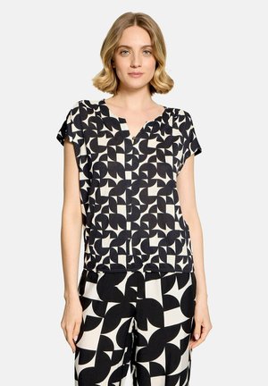 Vrouw met kort blond haar draagt een bijpassende zwart-wit geometrisch bedrukte top en broek, staand tegen een effen lichte achtergrond.