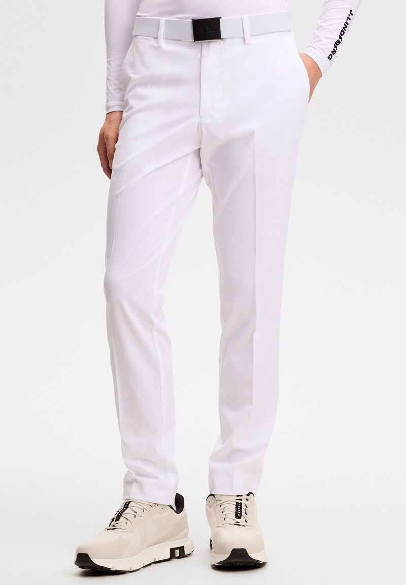 Personne portant un pantalon blanc sur mesure, des baskets beiges et une ceinture blanche, debout avec une main dans la poche devant un fond uni.