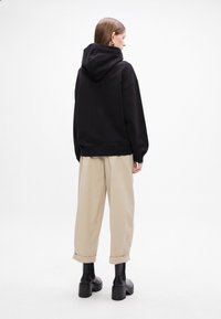 Felpa nera oversize con cappuccio e orlo a coste, abbinata a pantaloni cropped beige. Modella con stivali neri alla caviglia, vista da dietro.