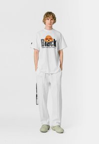 Camiseta blanca con un diseño gráfico de un oso y la palabra "Black" en negrita; combinada con pantalones blancos y zapatos de slip-on gruesos en verde claro.