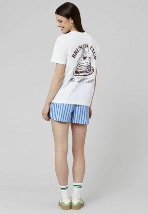 T-shirt blanc avec un graphique "BRUNCH CLUB" représentant un ours et un gâteau, associé à un short rayé bleu et blanc et des chaussures à accents verts.