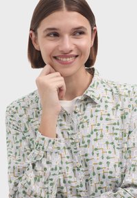 Camisa blanca de botones con un patrón de verduras verdes y amarillas. La tela es ligera con un acabado texturizado, lo que proporciona un aspecto casual.