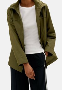 Oliwkowa wodoodporna kurtka z zamkiem błyskawicznym z przodu, regulowanym kapturem i bocznymi kieszeniami, w połączeniu z białą koszulą i czarnymi spodniami.