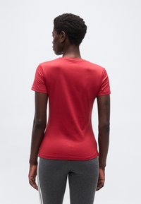 Άτομο που στέκεται γυρισμένο με την πλάτη, φορώντας στενό κόκκινο κοντομάνικο μπλουζάκι και γκρι κολάν με λευκές ρίγες στο πλάι, μπροστά σε απλό φόντο.