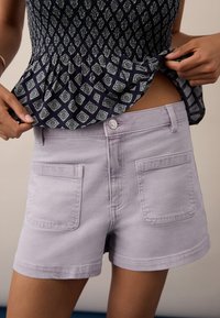 Pantaloni corti lavanda chiaro in denim, con chiusura a bottone, tasche anteriori e posteriori e una consistenza liscia. Abbinati a una maglietta stampata.