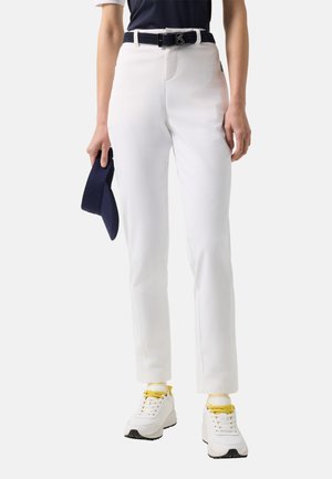 Personne portant un pantalon blanc à jambes droites, une ceinture noire avec une boucle argentée, des baskets blanches avec des lacets jaunes, tenant une casquette bleu marine dans la main gauche.