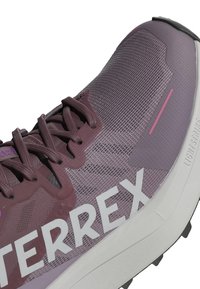 Zapatilla deportiva morada con parte superior de malla, logotipo "TERREX" en blanco en contraste, superficie texturizada y amortiguación Lightstrike en la suela.