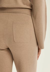 Beżowe dzianinowe spodnie z teksturowanym pasem i jedną łatką z tyłu, podkreślające gładkie wykończenie materiału.