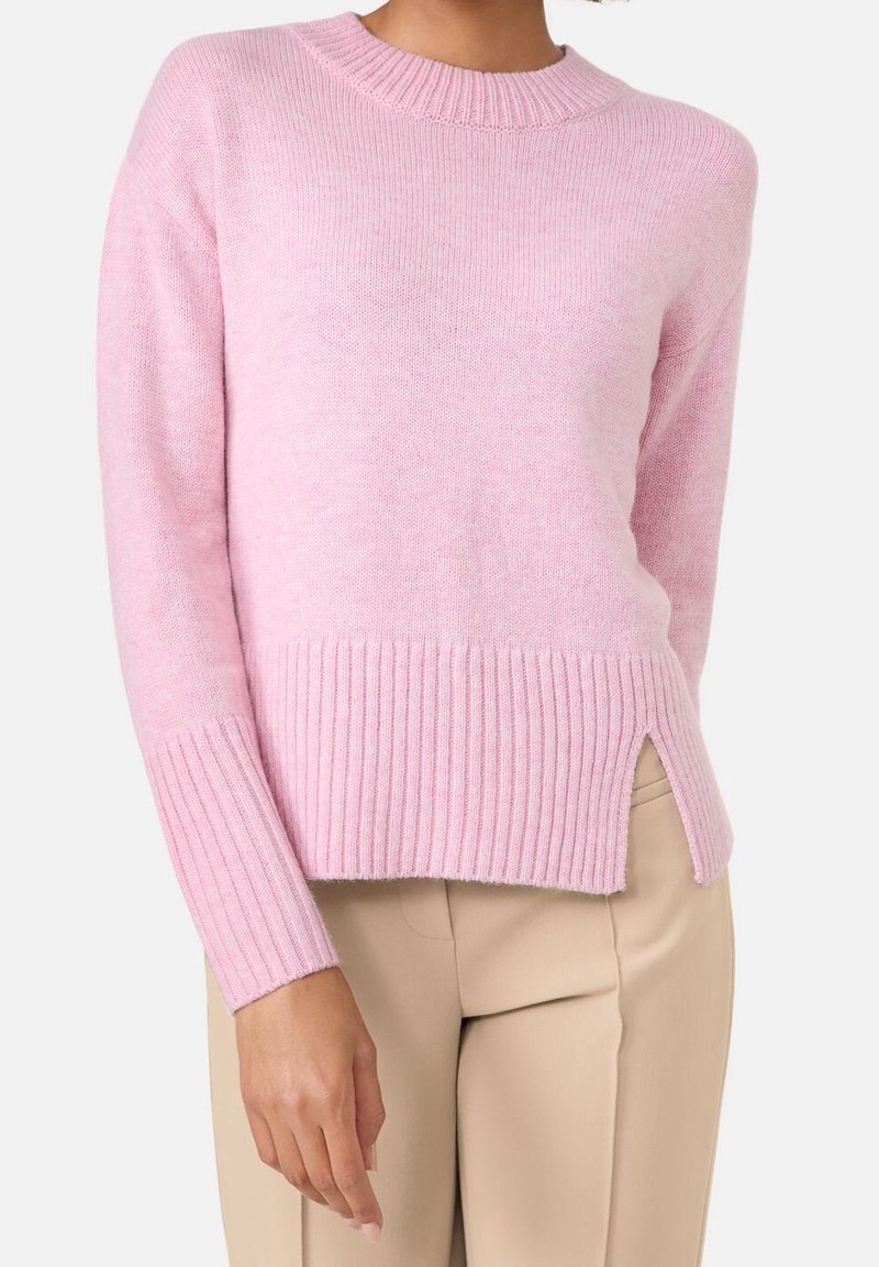 Femme portant un pull côtelé rose pâle à col rond associé à un pantalon beige, avec une fente latérale sur l'ourlet du pull.