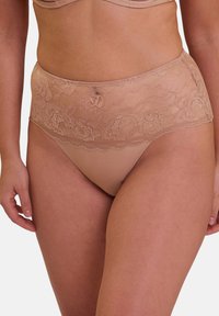Femme portant de la lingerie en dentelle haute taille beige et un soutien-gorge assorti, montrant le ventre et le haut des cuisses sur fond blanc.