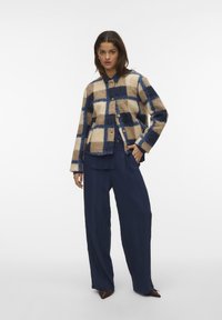 Wollen jas in een geruit patroon van beige en marineblauw, met een ronde hals, knoopsluiting en lange mouwen. Gecombineerd met marineblauwe broek.