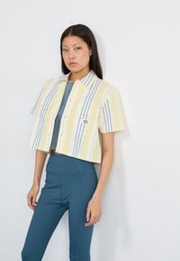 Beskyttende skjorte med gule og blå striper, korte ermer og to frontlommer. Brukt over en tettsittende blå topp og høy midje tealfargede leggings.