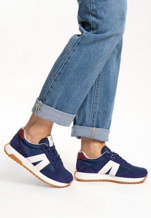 Person wearing cuffed blue jeans and navy blue sneakers with white accents and gum rubber soles, standing on a white background.