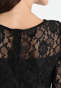 Robe en dentelle noire avec un motif floral, présentant un dos transparent et une fermeture éclair dissimulée au niveau du col. Détails en tissu texturé visibles.