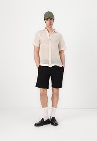 Lichtbeige opengehaakte shirt met korte mouwen, zwarte shorts, grijze pet, witte sokken en zwarte schoenen. Eenvoudig ontwerp met knoopsluiting.