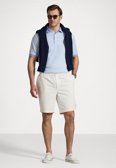 Polo texturat de culoare albastră deschisă, vestă din tricot navy, pantaloni scurți albi și adidași albi. Accesoriu: ochelari de soare negri. Outfit casual clasic.