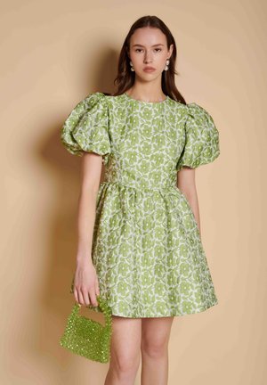 Jonge vrouw draagt een groene bloemenjurk met pofmouwen, houdt een kleine groene handtas met kralen vast, staand tegen een beige achtergrond.