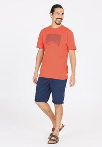 Herrarnas kortärmade, orange t-shirt med svart våg grafisk design, i kombination med blå shorts och bruna sandaler med spänne.