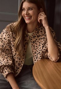 Leopardprint bomberjakke i beige stof med lynlås foran og afslappet pasform. Bæres over en lys grøn top med denimjeans.