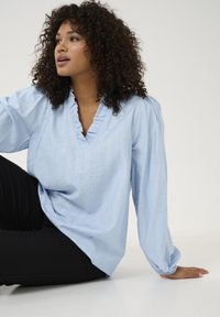 Blouse bleu clair avec une coupe ample, col en V et col froncé, tissu texturé, manches longues volumineuses, portée avec un pantalon noir.
