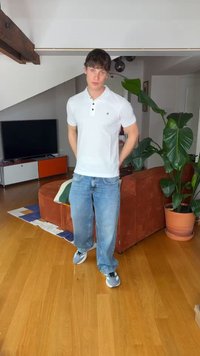 Młody mężczyzna w białej koszulce polo, luźnych niebieskich dżinsach i szarych tenisówkach stoi w nowoczesnym salonie z brązową sofą i dużą rośliną doniczkową.