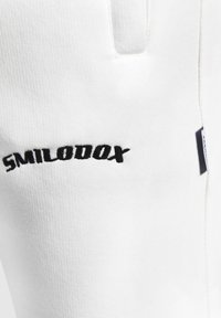 Hvide bomuldsjoggers med en tekstureret overflade, som har det sorte broderede logo "SMILODOX" på siden samt en skjult lomme. 