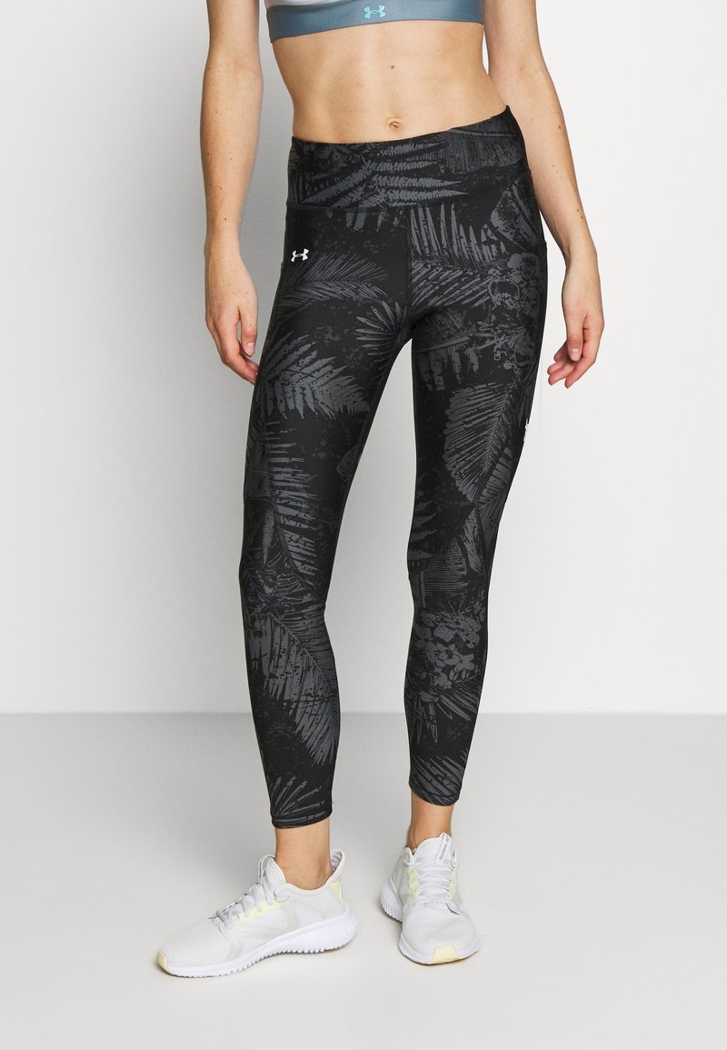 Femme portant des leggings noirs à motifs, un soutien-gorge de sport gris et des chaussures de sport blanches, debout devant un fond clair uni.