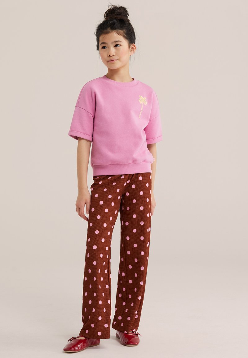 Jeune fille aux cheveux foncés en chignon, portant un sweat-shirt rose à manches courtes avec un palmier et un pantalon marron à pois roses.