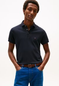 Vyras dėvi juodą trumpomis rankovėmis polo marškinėlį, įkištą į mėlynus džinsus su rudu diržu, stovi kišenėse laikydamas rankas priešais baltą foną.