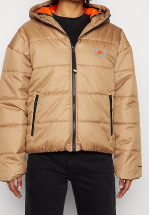 Veste matelassée beige avec fermeture éclair devant, poches zippées noires, doublure de capuche orange et petit patch logo sur la poitrine portée sur des vêtements noirs.