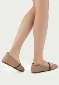 Beige tygskor med ribbad textur, ett brunt accentband och en rund metalldetalj. Varumärket "GOBI" är synligt på hälen.