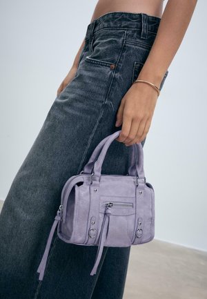 Kvinde iført slidte sorte jeans holder en lille lavendel håndtaske i håndtagene, med et guldarmbånd på sit håndled.
