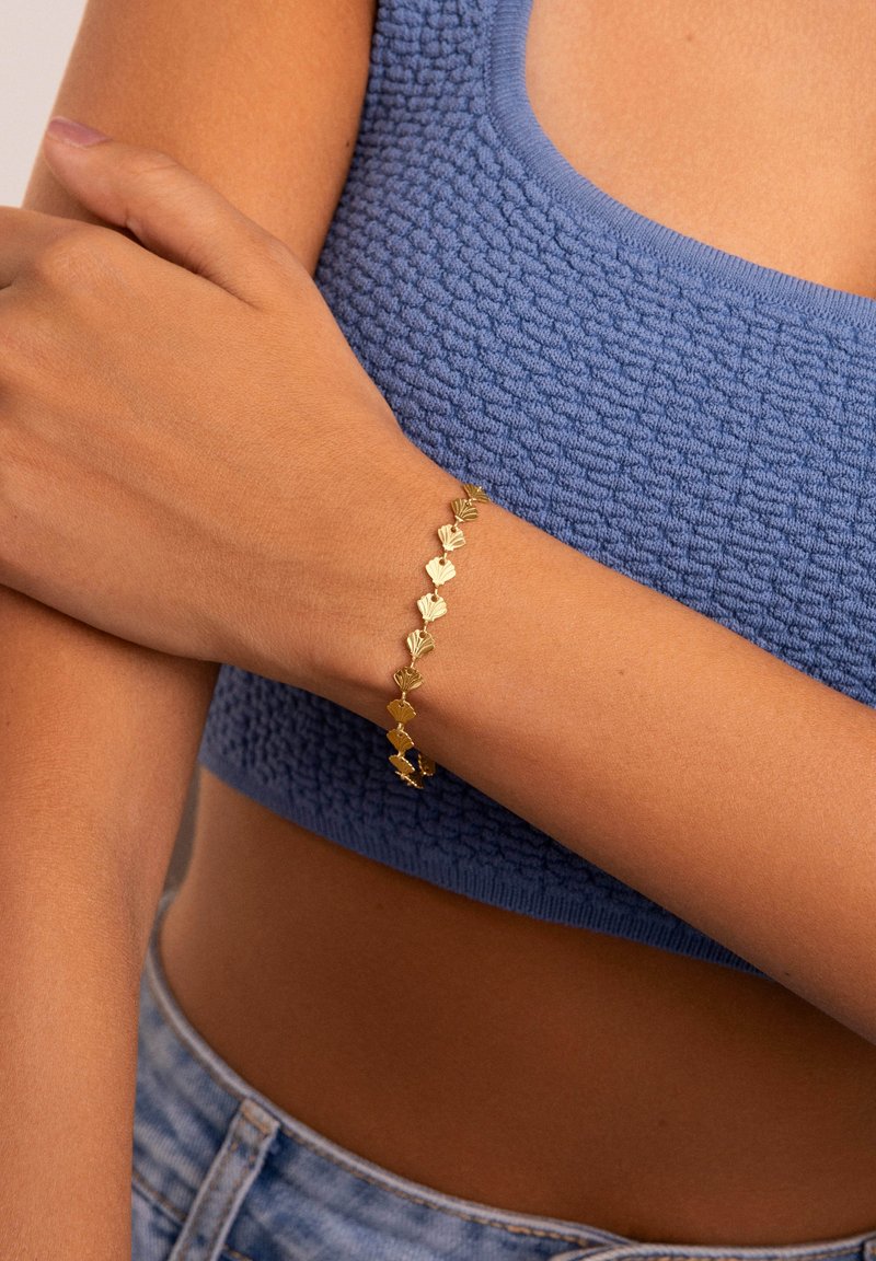 Bracciale d'oro con una serie di forme geometriche e una finitura lucida, indossato su un polso contro una maglietta blu strutturata.