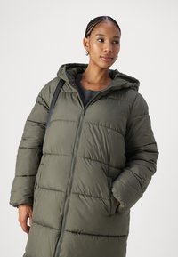 Cappotto imbottito di un verde oliva scuro, con chiusura frontale con zip, cappuccio e ampie tasche. Il design presenta dettagli trapuntati orizzontali e una texture liscia.