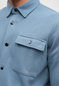 Svetle modrá košile s texturovanou tkaninou, s límečkem, černými druky a náprsní kapsou. Hladký povrch s jemným detailem.