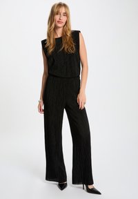 Zwarte jumpsuit met een mouwloos ontwerp, geplooide textuur, wijde pijpen en een aansluitende taille. Gecombineerd met zwarte puntige hakken.