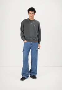 Felpa grigia con polsini e orlo a costine, jeans cargo blu con tasche, scarpe nere e una collana d'argento. Design semplice e casual.