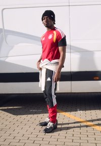 Sarkans un melns Adidas sporta tērps ar trim svītrām, melns cepure un balts džemperis sasiets ap vidu, stāv uz strukturētas bruģētas virsmas.