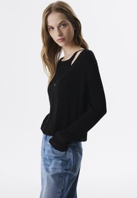 Pull en tricot noir côtelé à manches longues, avec un col rond et un petit découpe décoratif. Associé à un jean en denim bleu clair.