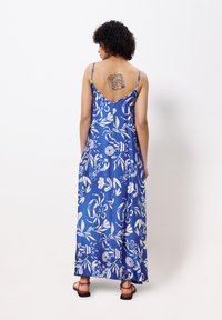 Lang blauwe jurk met wit bloemenpatroon, spaghettibandjes, lage rug en een vloeiende ontwerp. Draagt rode sandalen en heeft een zichtbare tatoeage.