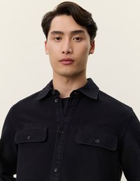 Μαύρο πουκάμισο από χοντρό ύφασμα με κουμπιά στο μπροστινό μέρος, δύο τσέπες στο στήθος και παραδοσιακό γιακά, που αναδεικνύει μια υφή υφάσματος.