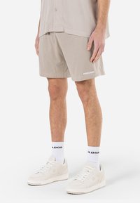 Shorts en maille beige avec une taille élastique, arborant un logo blanc en bas, associés à des baskets blanches et des chaussettes avec logo.