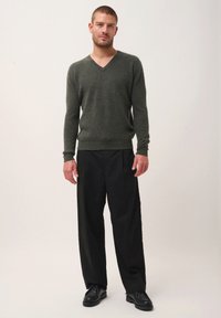 Donkergroene wollen trui met V-hals en geribbelde boorden, gecombineerd met losse zwarte broek en zwarte schoenen. Model staat op een effen achtergrond.