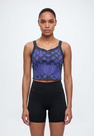 Une femme présente un crop top violet à motifs avec des bretelles noires et un short de cyclisme noir sur un fond blanc uni.