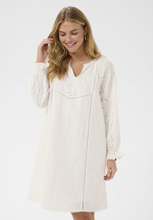 Donna sorridente con capelli biondi lunghi che indossa un vestito bianco ampio con motivi traforati e ricamati, posando davanti a uno sfondo semplice.