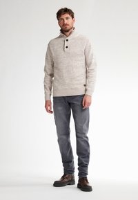 Pullover in maglia grigia chiaro con colletto alto, caratterizzato da due bottoni scuri e un bordo a costine, abbinato a jeans scuri e stivali marroni con lacci.